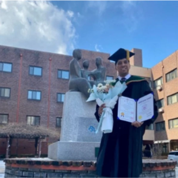 A graduate in a cap and gown smiles while holding a diploma and bouquet in front of a university statue