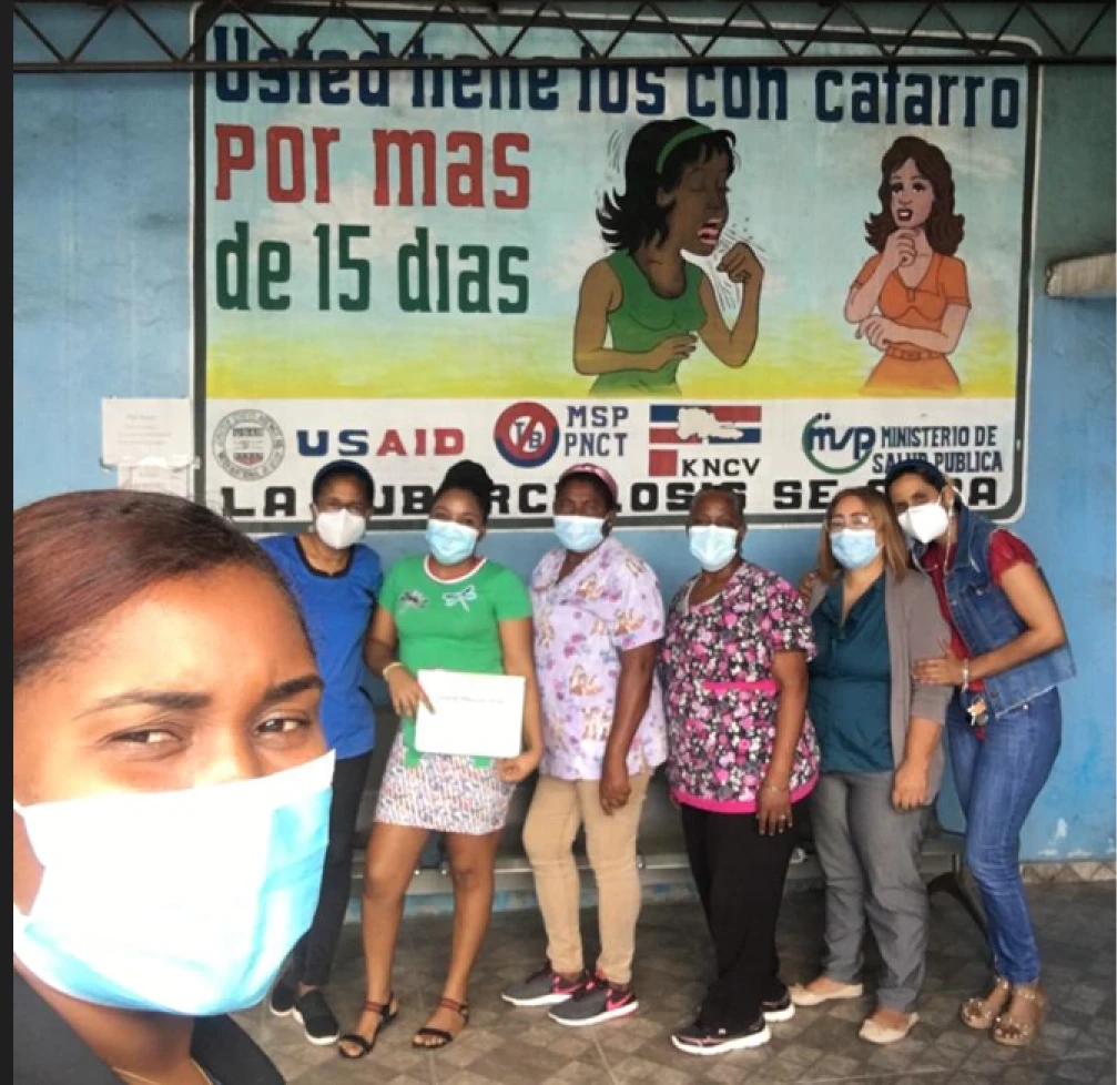 A group of women wearing face masks pose in front of a mural with Spanish text about persistent cough symptoms and tuberculosis awareness