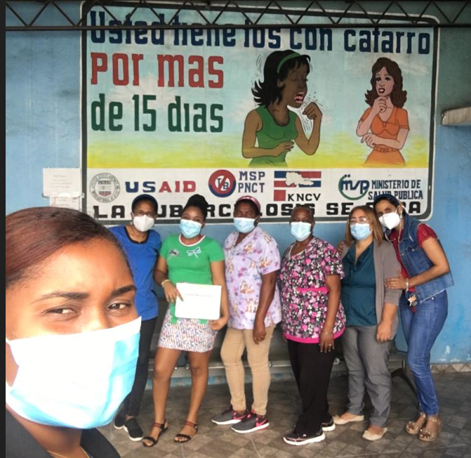 A group of women wearing face masks pose in front of a mural with Spanish text about persistent cough symptoms and tuberculosis awareness